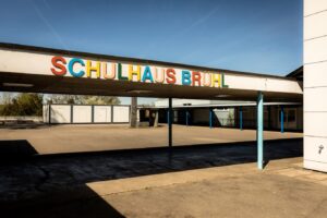 Entrance to Schulhaus Brühl: a low, flat-roofed school building with a colorful, multicolored sign and blue support columns under a sheltered walkway.