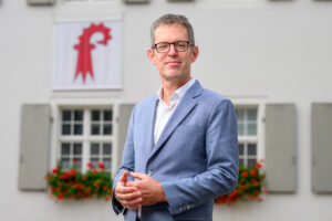 Smiling man in a light-blue suit posing outdoors in front of a white building with a red emblem banner and flower boxes.
