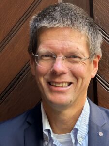Smiling man with short gray hair and round glasses, wearing a navy blazer and striped shirt in front of a wooden door backdrop.