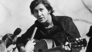 Male musician with an acoustic guitar being interviewed by reporters; a handheld microphone is held close to him in a black-and-white photo.