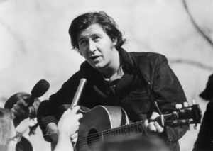 Male musician with an acoustic guitar being interviewed by reporters; a handheld microphone is held close to him in a black-and-white photo.