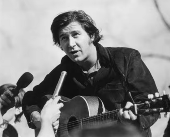 Male musician with an acoustic guitar being interviewed by reporters; a handheld microphone is held close to him in a black-and-white photo.