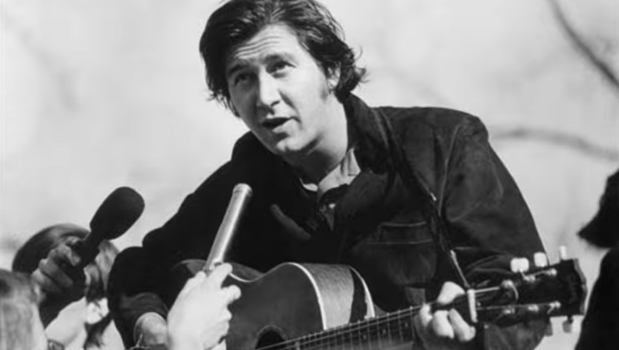 Male musician with an acoustic guitar being interviewed by reporters; a handheld microphone is held close to him in a black-and-white photo.