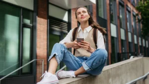 Young woman with a backpack sits on a concrete railing outside a brick building, looking up while using her phone.