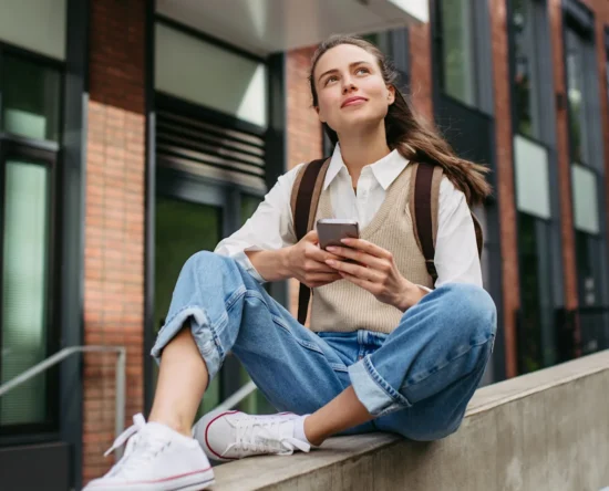 Young woman with a backpack sits on a concrete railing outside a brick building, looking up while using her phone.