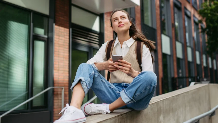 Young woman with a backpack sits on a concrete railing outside a brick building, looking up while using her phone.