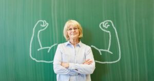 Older woman with crossed arms in front of a green chalkboard, framed by large chalk-drawn flexed arms behind her.