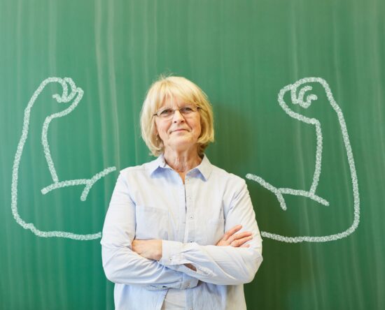 Older woman with crossed arms in front of a green chalkboard, framed by large chalk-drawn flexed arms behind her.