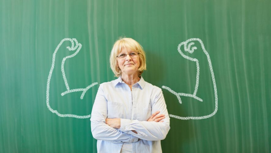 Older woman with crossed arms in front of a green chalkboard, framed by large chalk-drawn flexed arms behind her.
