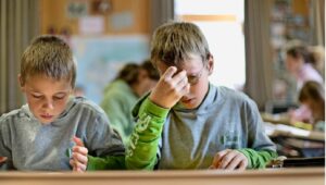 Two young boys sit at a desk in a classroom, focused on work, one adjusting his glasses with his hand.