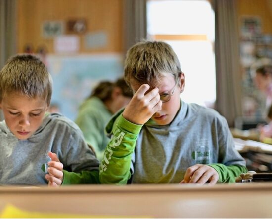 Two young boys sit at a desk in a classroom, focused on work, one adjusting his glasses with his hand.