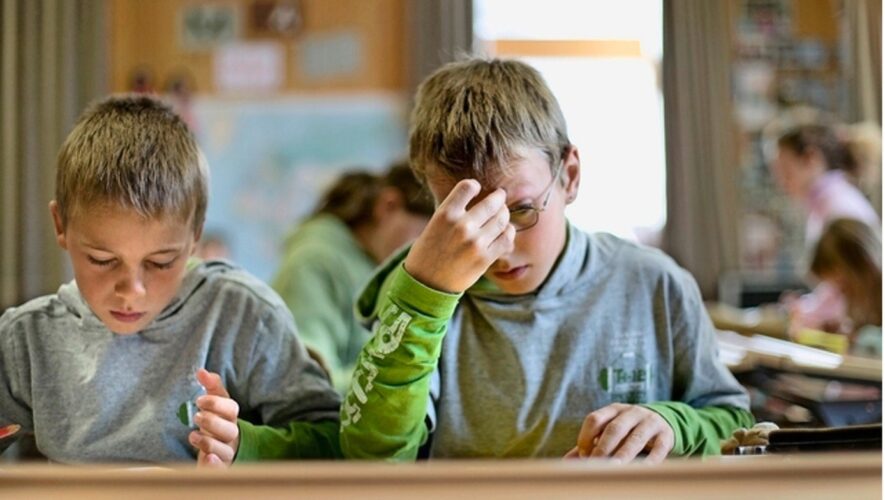 Two young boys sit at a desk in a classroom, focused on work, one adjusting his glasses with his hand.