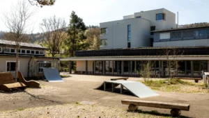 Outdoor courtyard of a modern building with wooden benches, a small skate ramp, and a planted area under clear sky