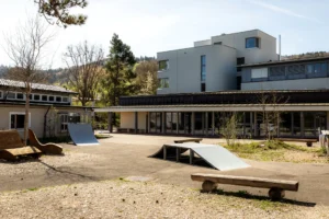 Outdoor courtyard of a modern building with wooden benches, a small skate ramp, and a planted area under clear sky