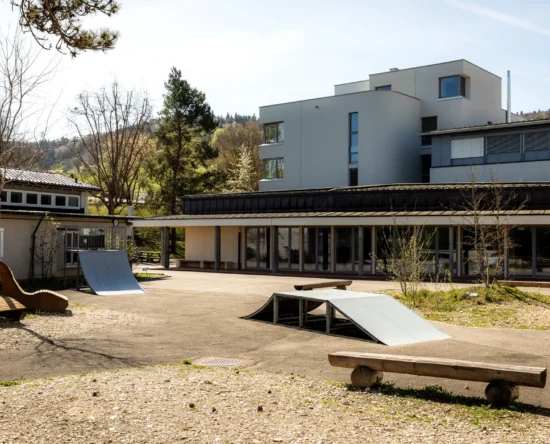 Outdoor courtyard of a modern building with wooden benches, a small skate ramp, and a planted area under clear sky