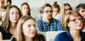 Classroom full of students listening attentively, centered on a man wearing glasses and a blue plaid shirt.