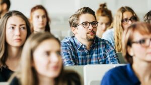 Classroom full of students listening attentively, centered on a man wearing glasses and a blue plaid shirt.