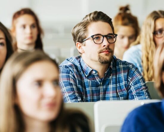 Classroom full of students listening attentively, centered on a man wearing glasses and a blue plaid shirt.