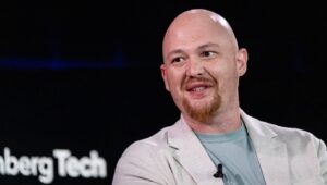 Bald man with a red goatee wearing a light blazer and teal shirt, speaking on stage with a clip-on mic.