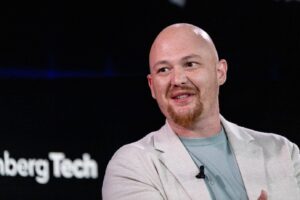 Bald man with a red goatee wearing a light blazer and teal shirt, speaking on stage with a clip-on mic.