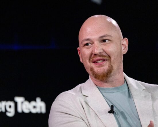 Bald man with a red goatee wearing a light blazer and teal shirt, speaking on stage with a clip-on mic.