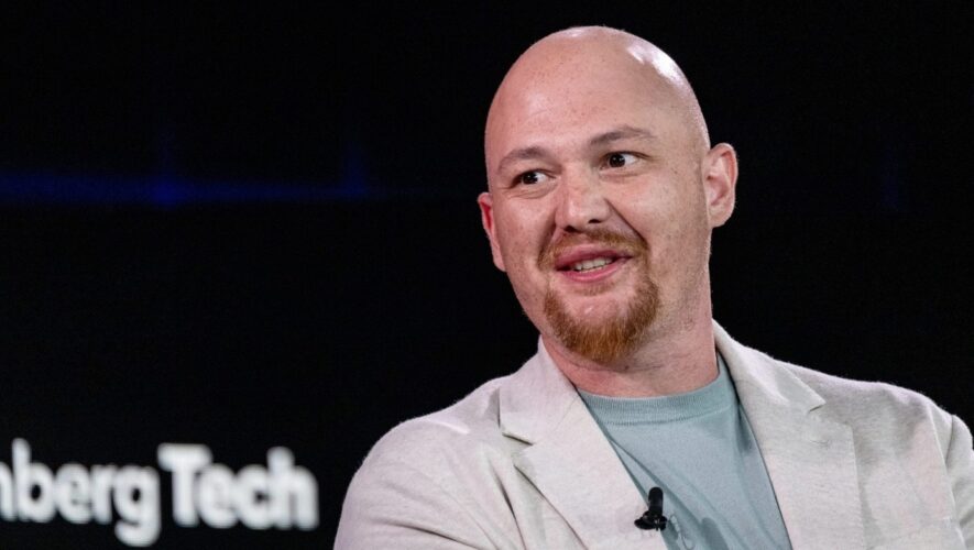Bald man with a red goatee wearing a light blazer and teal shirt, speaking on stage with a clip-on mic.