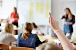 Raised hand of a student in a classroom, signaling to participate during a lesson; blurred classmates in the background.