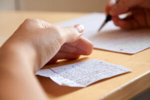 Close-up of a hand writing with a pencil on a lined sheet of paper on a wooden desk.