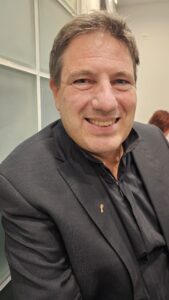 Smiling man in a dark blazer seated indoors, looking at the camera against a glass-paneled wall.