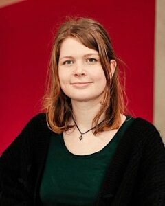 Portrait of a young woman with shoulder-length brown hair, wearing a green top and black cardigan, against a red background.