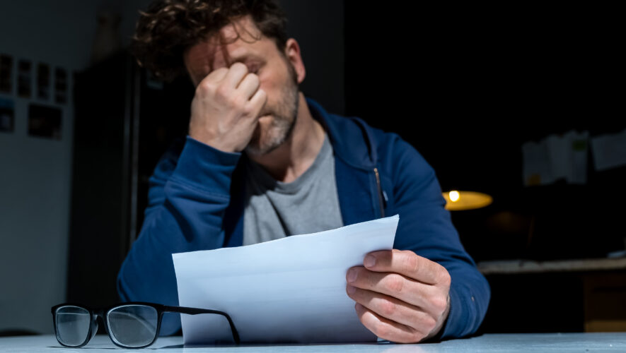 Man in a blue jacket sits at a desk, hand over his face, reading a stack of papers; glasses on the desk in the foreground.