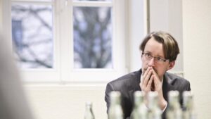 A man in a suit sits at a table in a meeting room, hands clasped near his mouth and a thoughtful expression.