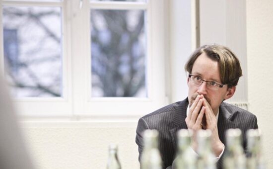 A man in a suit sits at a table in a meeting room, hands clasped near his mouth and a thoughtful expression.