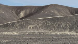 Desert landscape with brown hills and a row of ancient petroglyphs carved into the hillside, foreground rocky ground.
