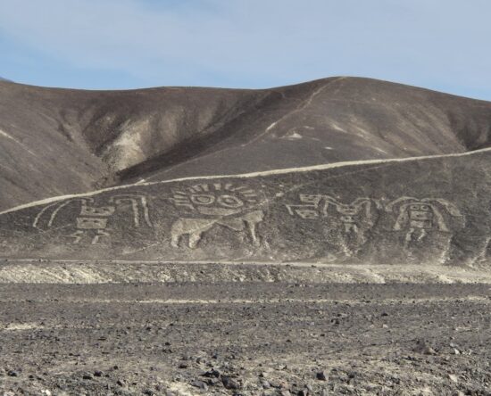 Desert landscape with brown hills and a row of ancient petroglyphs carved into the hillside, foreground rocky ground.