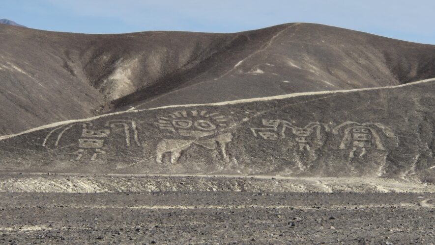 Desert landscape with brown hills and a row of ancient petroglyphs carved into the hillside, foreground rocky ground.
