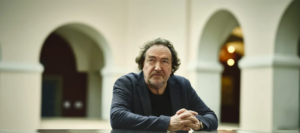 Man in a dark blazer sits at a table in a light arched courtyard, hands clasped, looking at the camera.