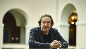 Man in a dark blazer sits at a table in a light arched courtyard, hands clasped, looking at the camera.