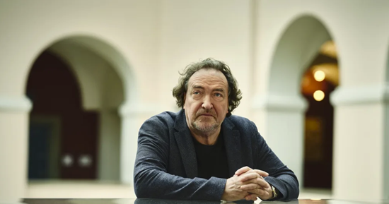 Man in a dark blazer sits at a table in a light arched courtyard, hands clasped, looking at the camera.