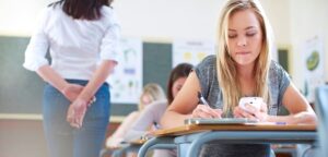 Student at a classroom desk writes with a pen while holding a smartphone.