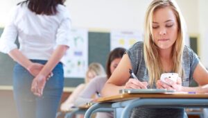 Student at a classroom desk writes with a pen while holding a smartphone.