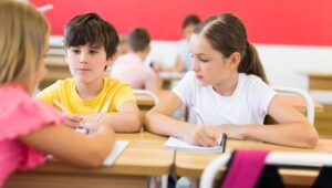 Two children sit at a shared classroom desk, writing in notebooks as a pink-clad classmate speaks nearby (blurred foreground).