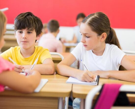Two children sit at a shared classroom desk, writing in notebooks as a pink-clad classmate speaks nearby (blurred foreground).