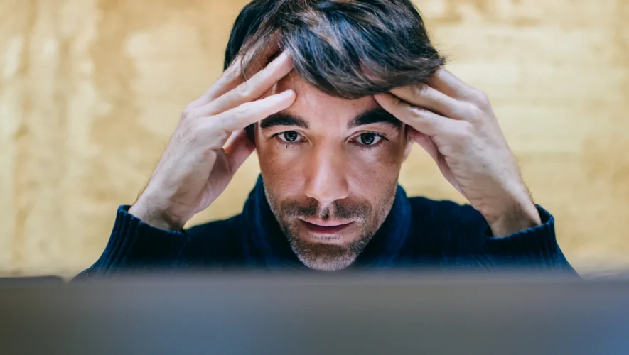 Man concentrates in front of a screen, hands pressed to temples as he looks at the monitor.