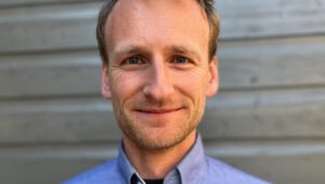 Close-up of a smiling man in a blue button-down shirt against a wooden wall background.
