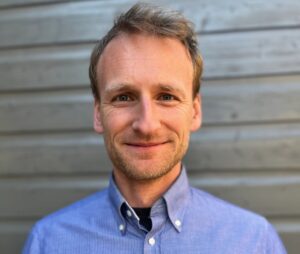 Close-up of a smiling man in a blue button-down shirt against a wooden wall background.