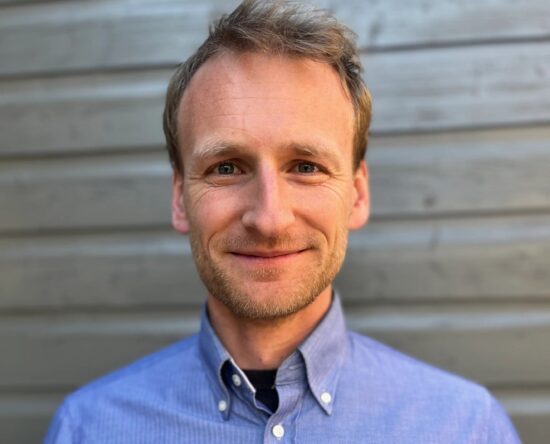 Close-up of a smiling man in a blue button-down shirt against a wooden wall background.