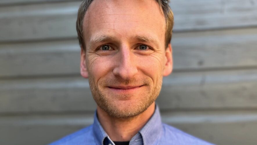 Close-up of a smiling man in a blue button-down shirt against a wooden wall background.