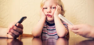 A young child in a striped shirt sits at a table with hands on cheeks, looking upset as two adults hold phones nearby.