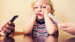 A young child in a striped shirt sits at a table with hands on cheeks, looking upset as two adults hold phones nearby.